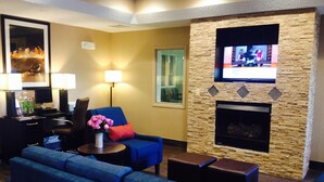 A hotel lobby with a stone fireplace, a television mounted on the wall, a blue sofa, a desk with a computer, and a lamp. - Comfort Inn Crystal Lake - Algonquin (Crystal Lake)