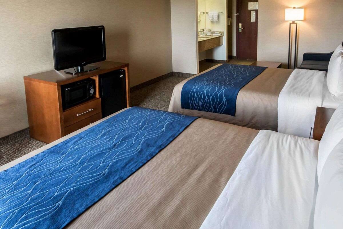 A hotel room with two beds, a TV on a wooden cabinet, a bathroom visible through an open door, and a floor lamp.