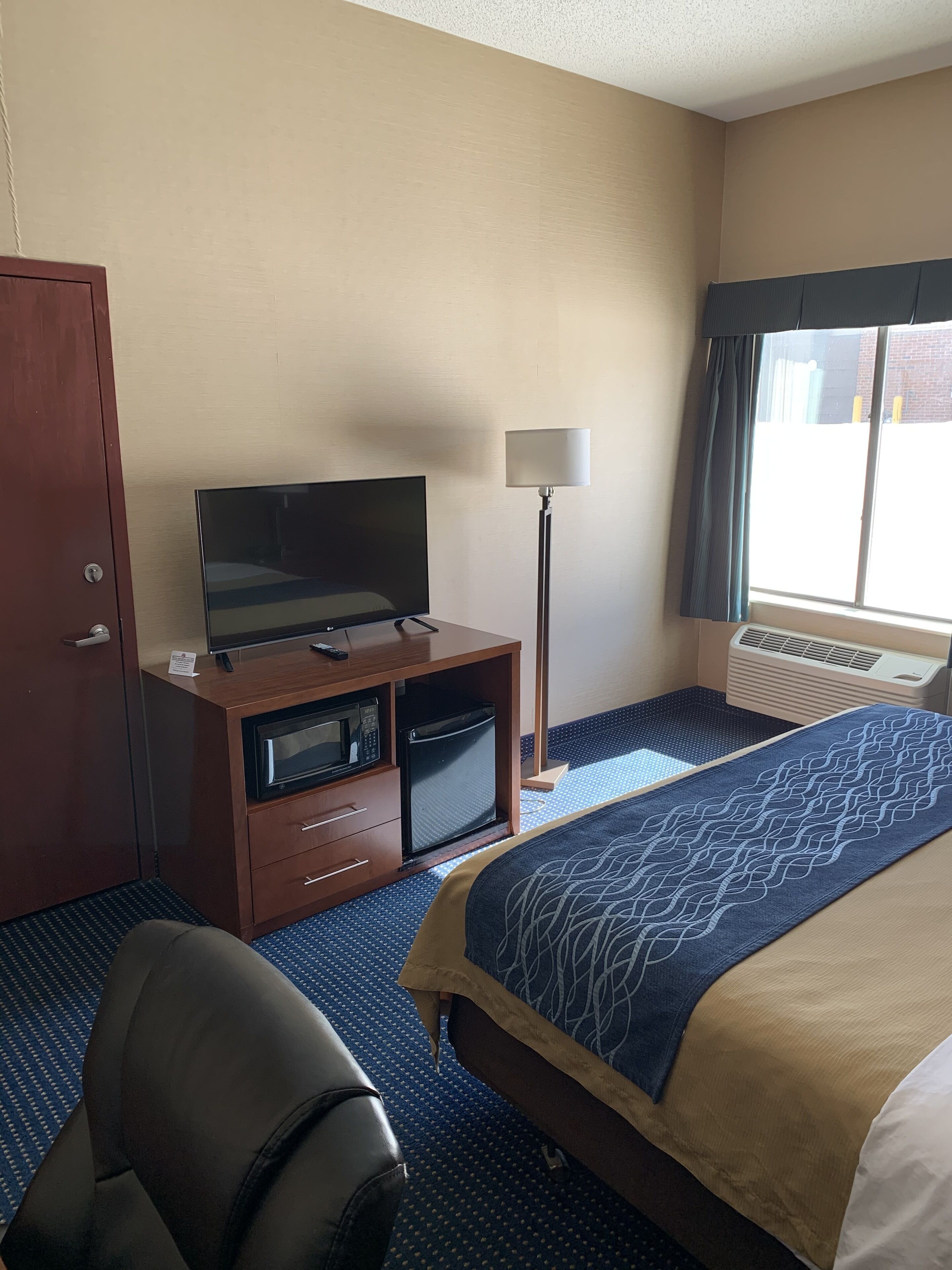 A hotel room with a bed, a desk with a television, a lamp, a chair, and a window with curtains.