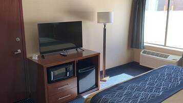 A hotel room with a bed, a desk with a television, a lamp, a chair, and a window with curtains.
