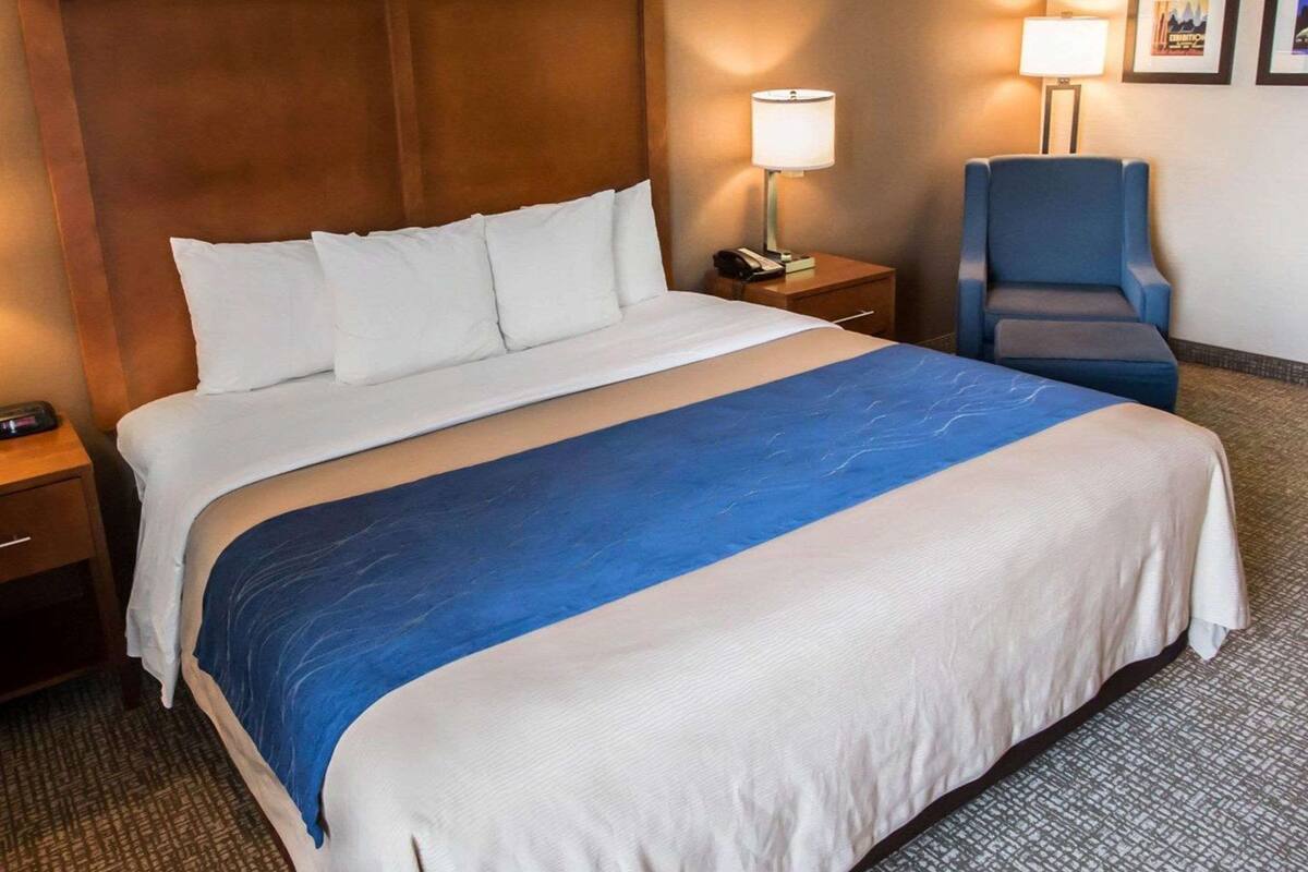 A hotel room with a large bed, a wooden headboard, a blue accent on the bedspread, a blue armchair, two bedside lamps, and framed pictures on the wall.