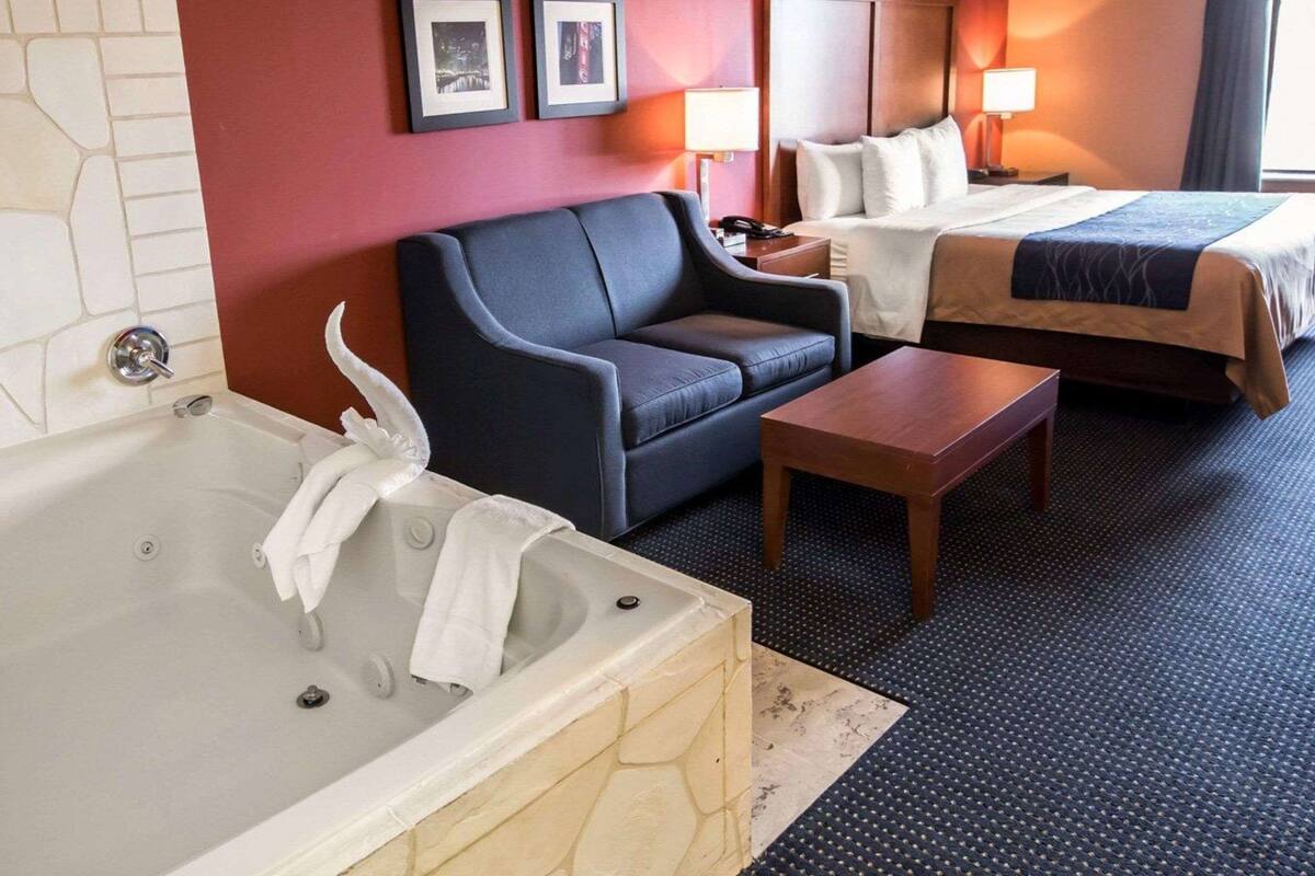 A hotel room with a bathtub, two beds, a sofa, a small table, and two wall-mounted lamps.