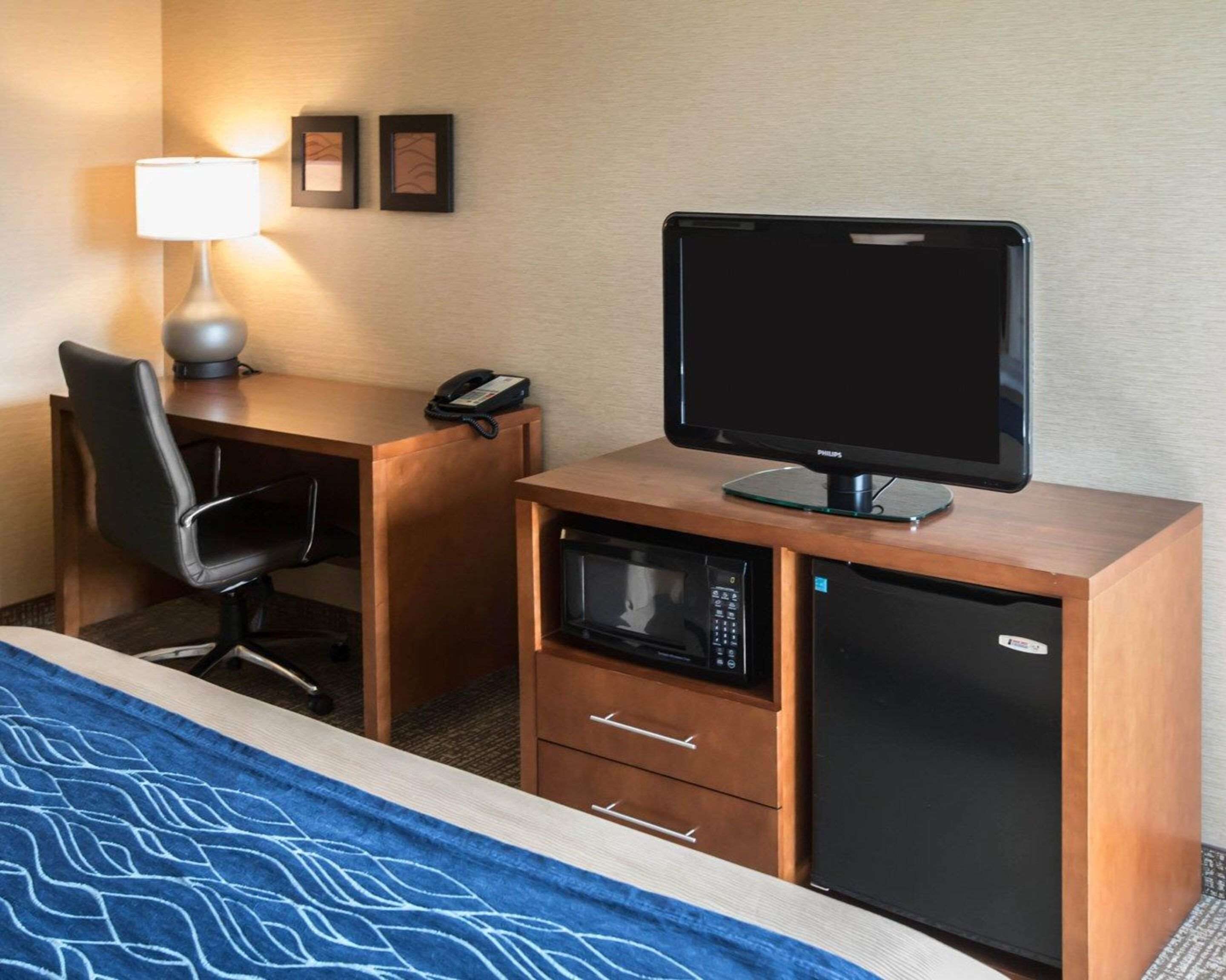 A hotel room with a bed, a desk with a computer monitor, a telephone, a microwave, and a lamp.