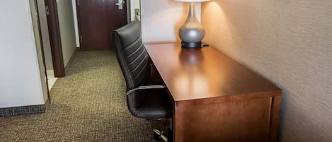 A hotel room with a wooden desk, a black office chair, a lamp, and a carpeted floor.