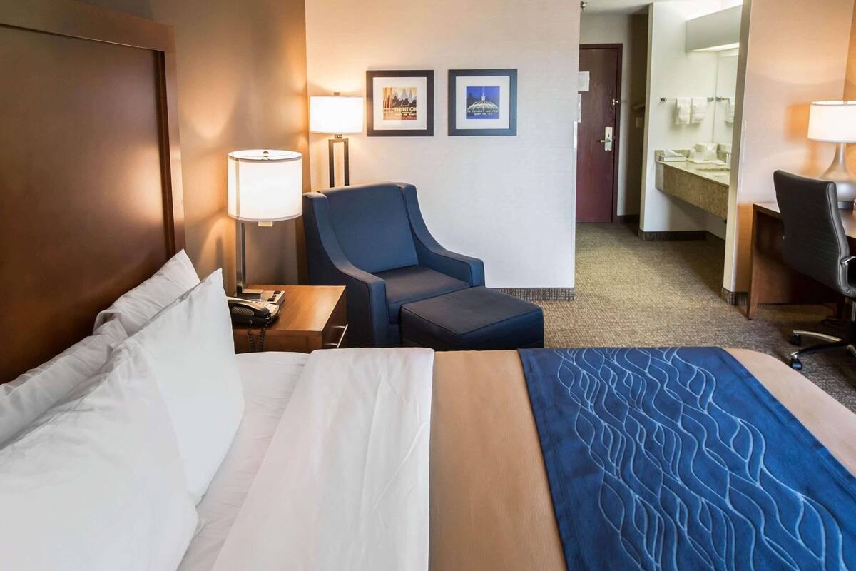 A hotel room with a bed, a desk with a lamp, a chair, and a bathroom visible in the background.