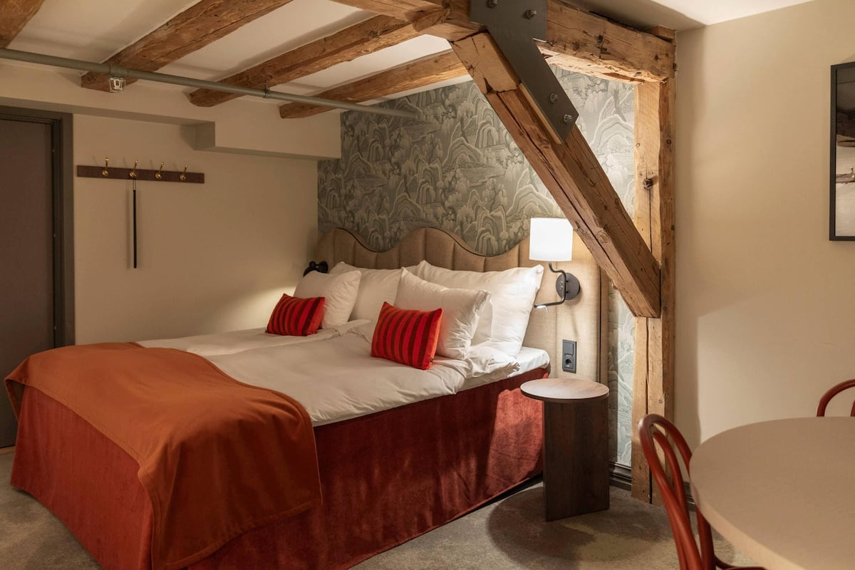 A bedroom with a bed, a bedside table, a lamp, a coat rack, and a window with a view.
