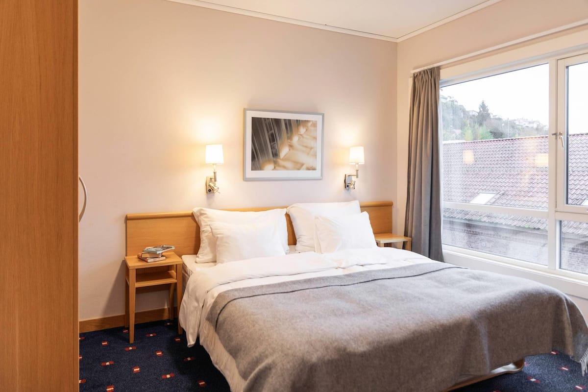 A hotel room with a bed, bedside lamps, a nightstand, a framed picture on the wall, and a window with a view of buildings.
