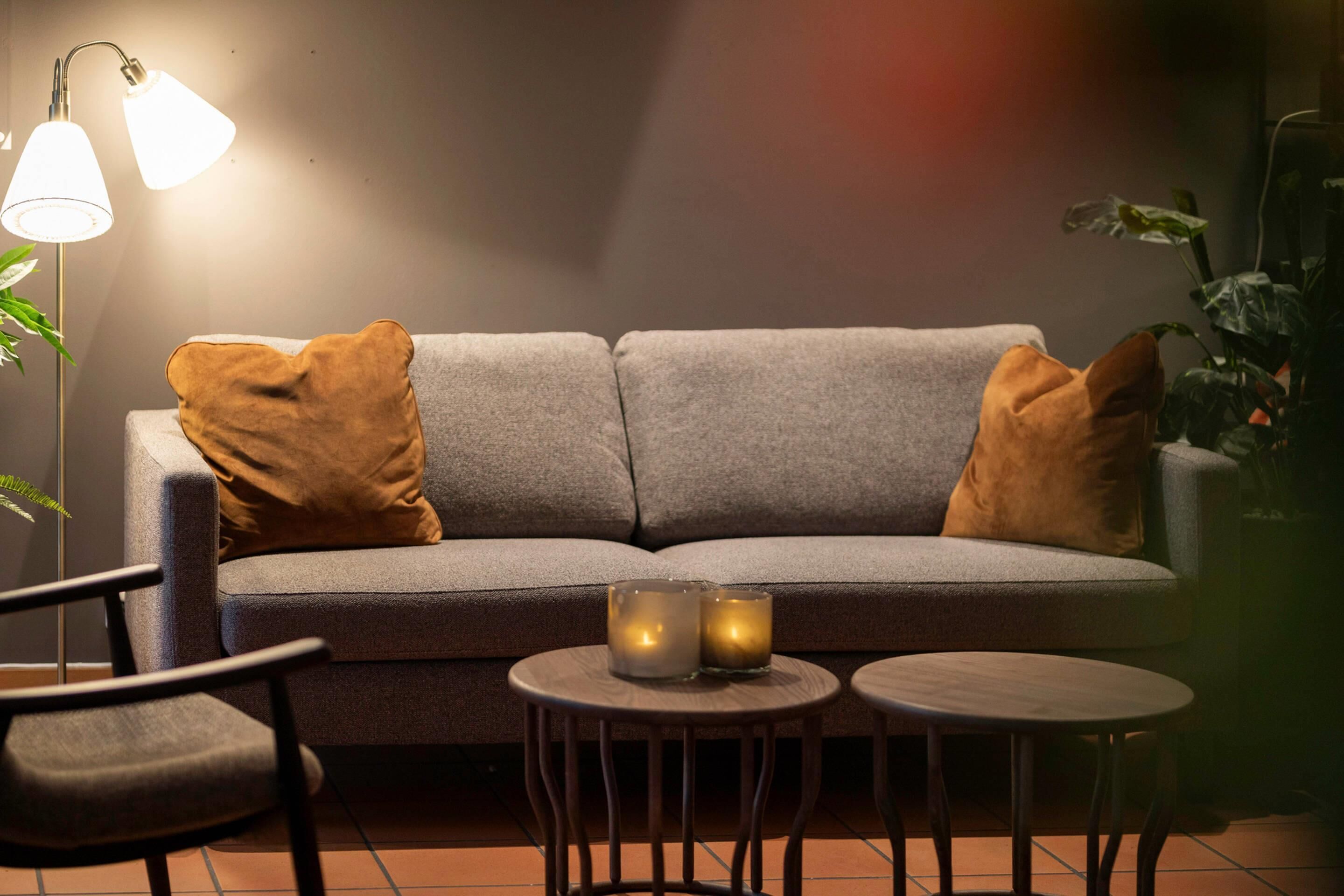 A grey sofa with two brown cushions, two small round tables, a floor lamp, and a potted plant.