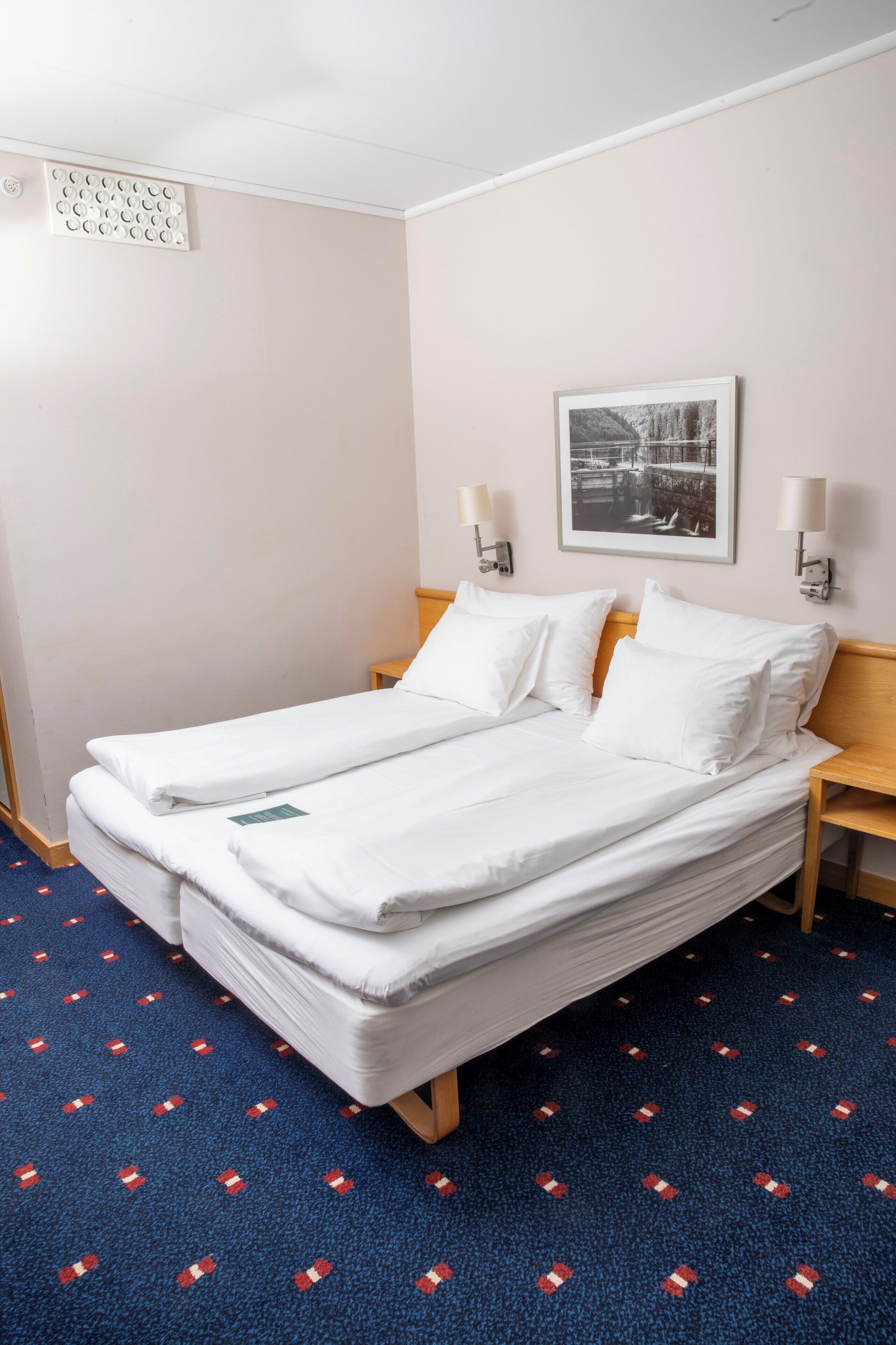Una cama doble con sábanas blancas, dos mesitas de noche con lámparas, un cuadro en la pared y una alfombra estampada.