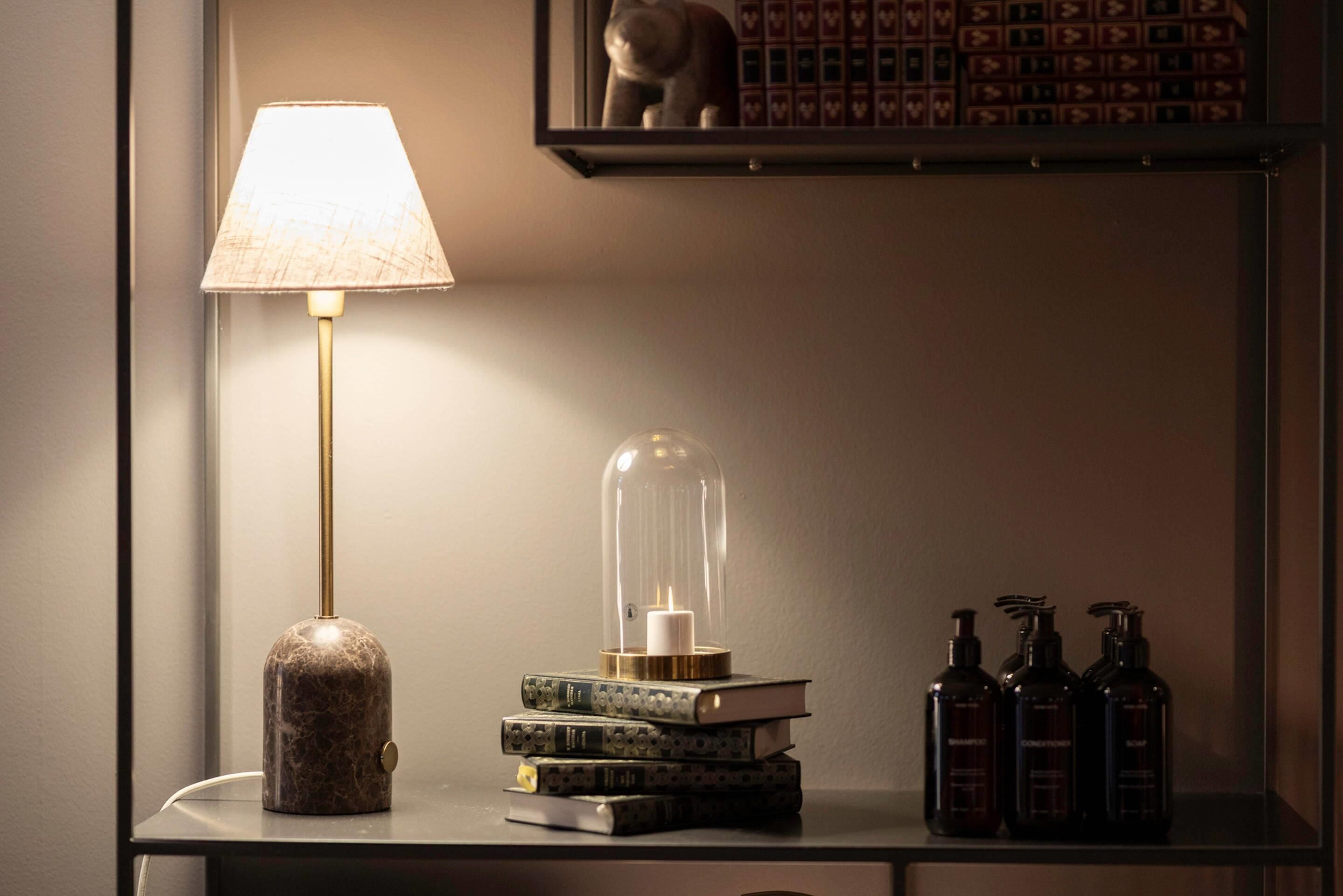 A bedside table with a lamp, a glass dome candle holder, stacked books, and three bottles.