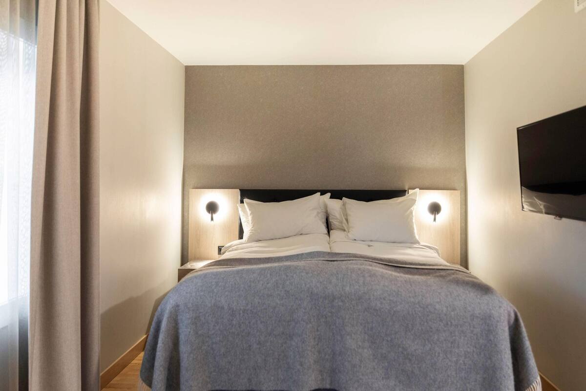 A hotel room with a bed, two bedside lamps, a TV, and a window with curtains.