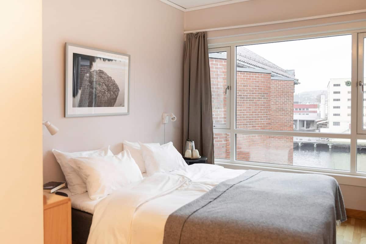 A hotel room with a bed, a nightstand, a framed picture on the wall, a window with a view of buildings, and a curtain.