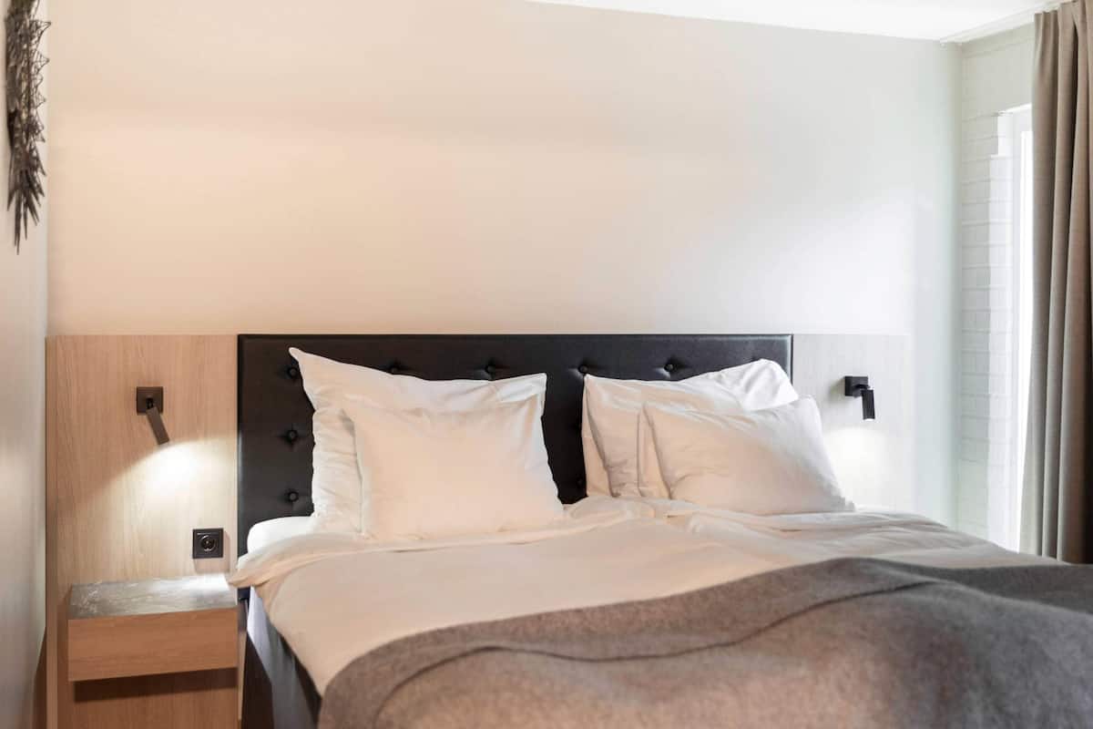A hotel room with a bed, a nightstand, a wall-mounted light, and a window with curtains.
