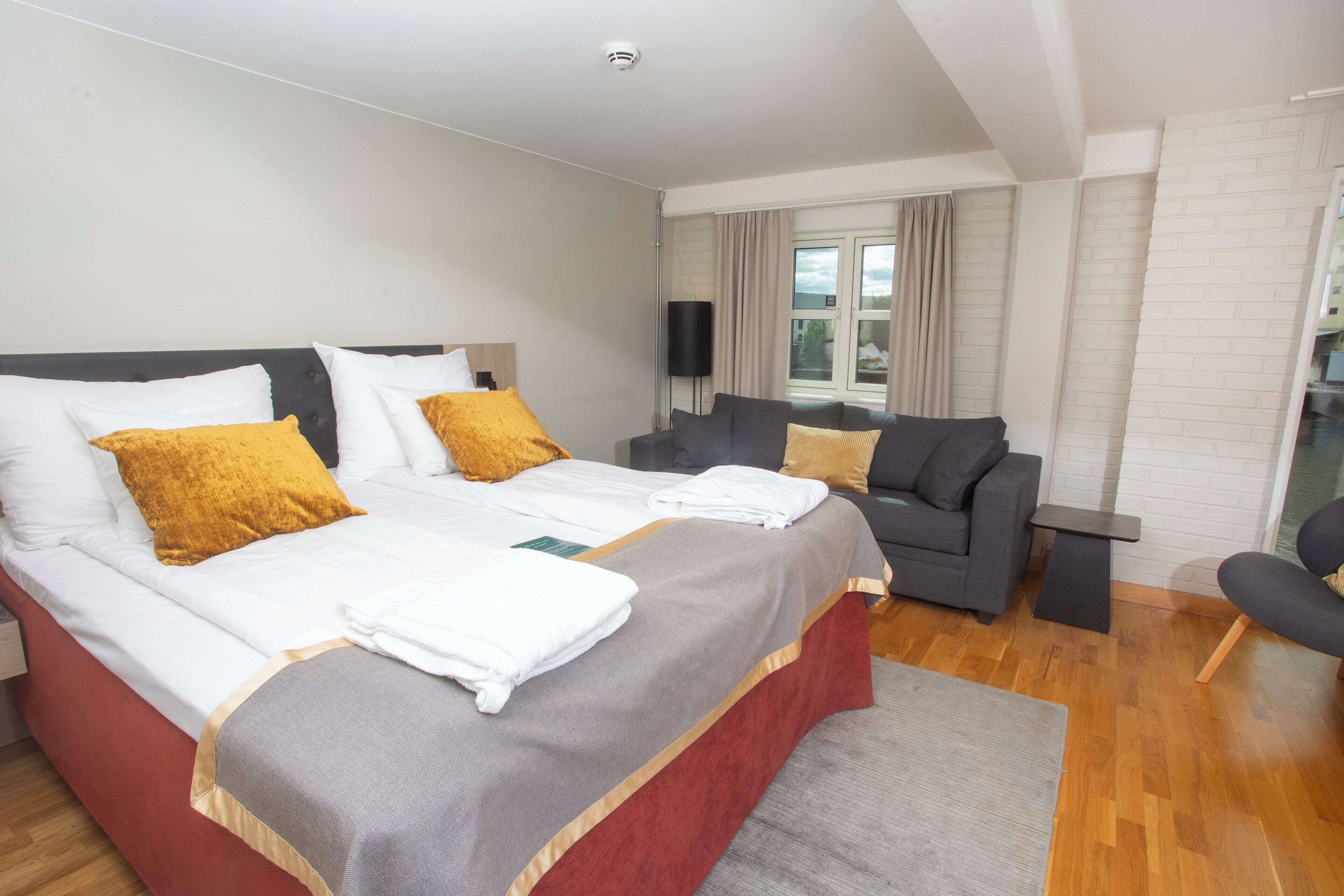 Et moderne hotelværelse med en stor seng, en grå sofa, et lille bord og udsigt til det fri gennem et vindue.