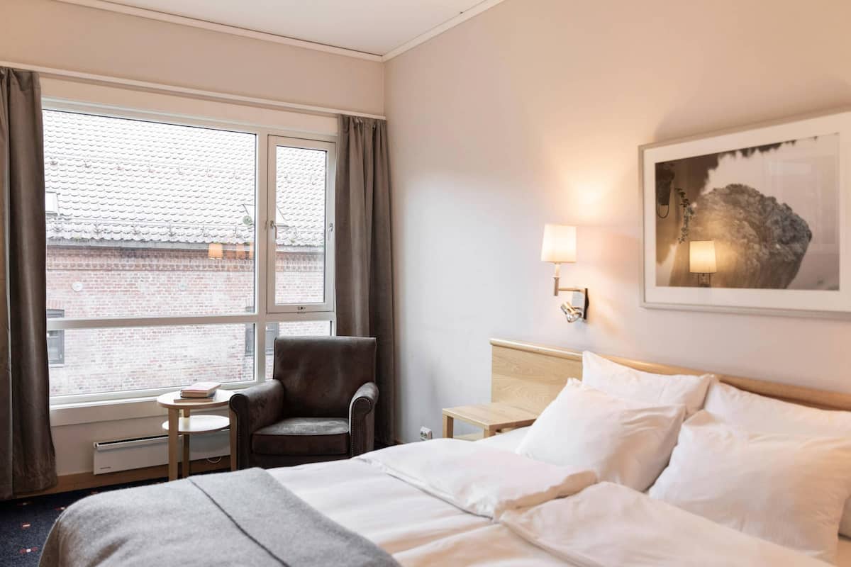 A hotel room with a bed, a chair, a small table, a lamp, and a window with curtains.