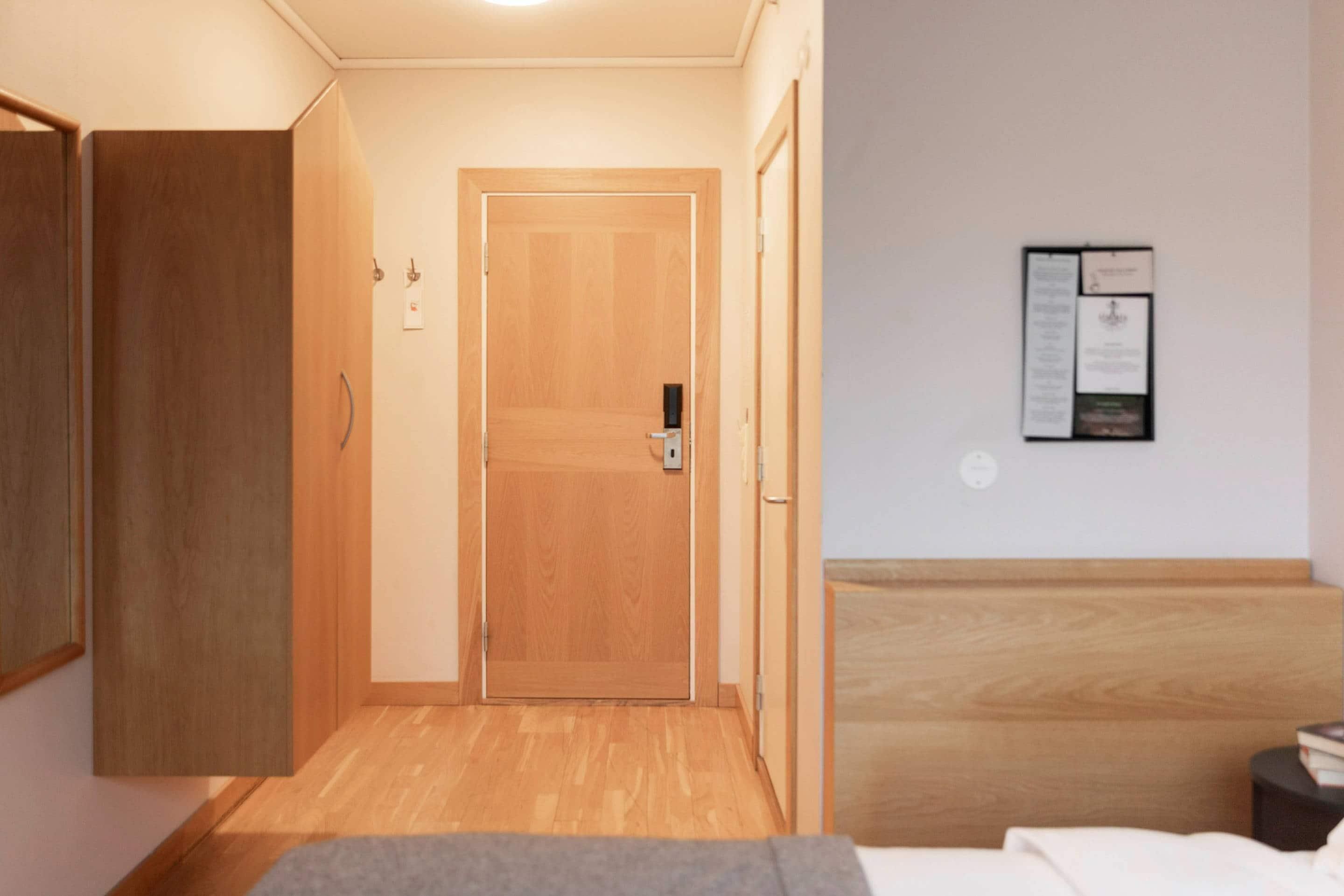Et hotelværelse med en trædør, en seng, et natbord og en opslagstavle på væggen.