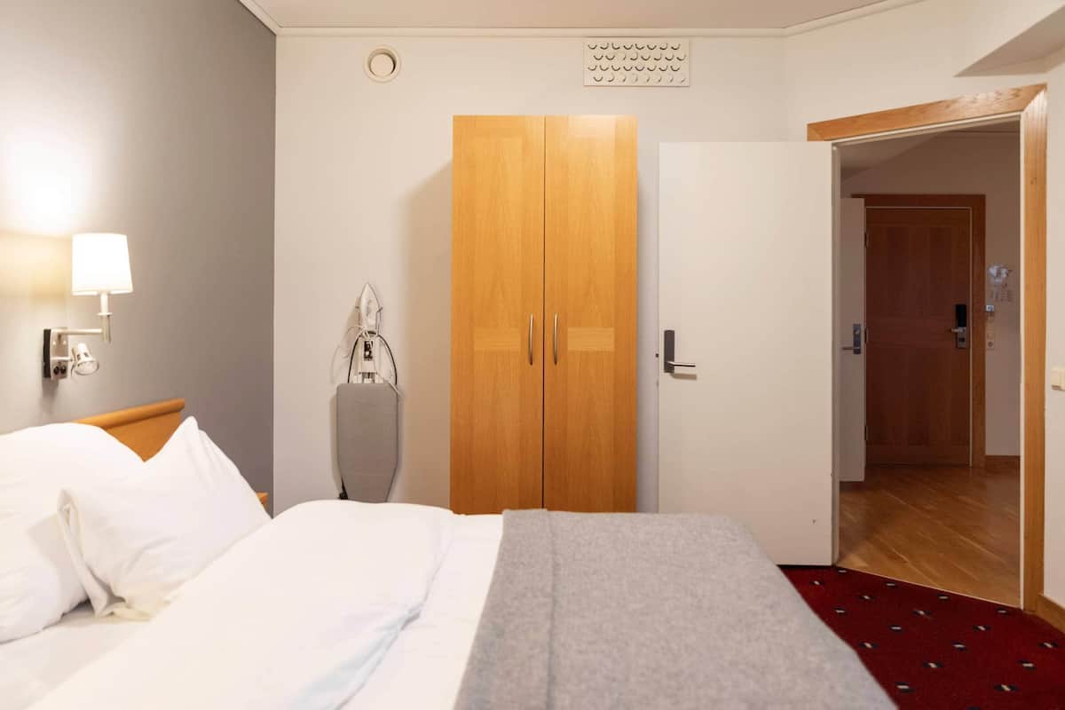 A hotel room with a bed, a wardrobe, a coat rack, a wall-mounted lamp, and a door leading to another room.