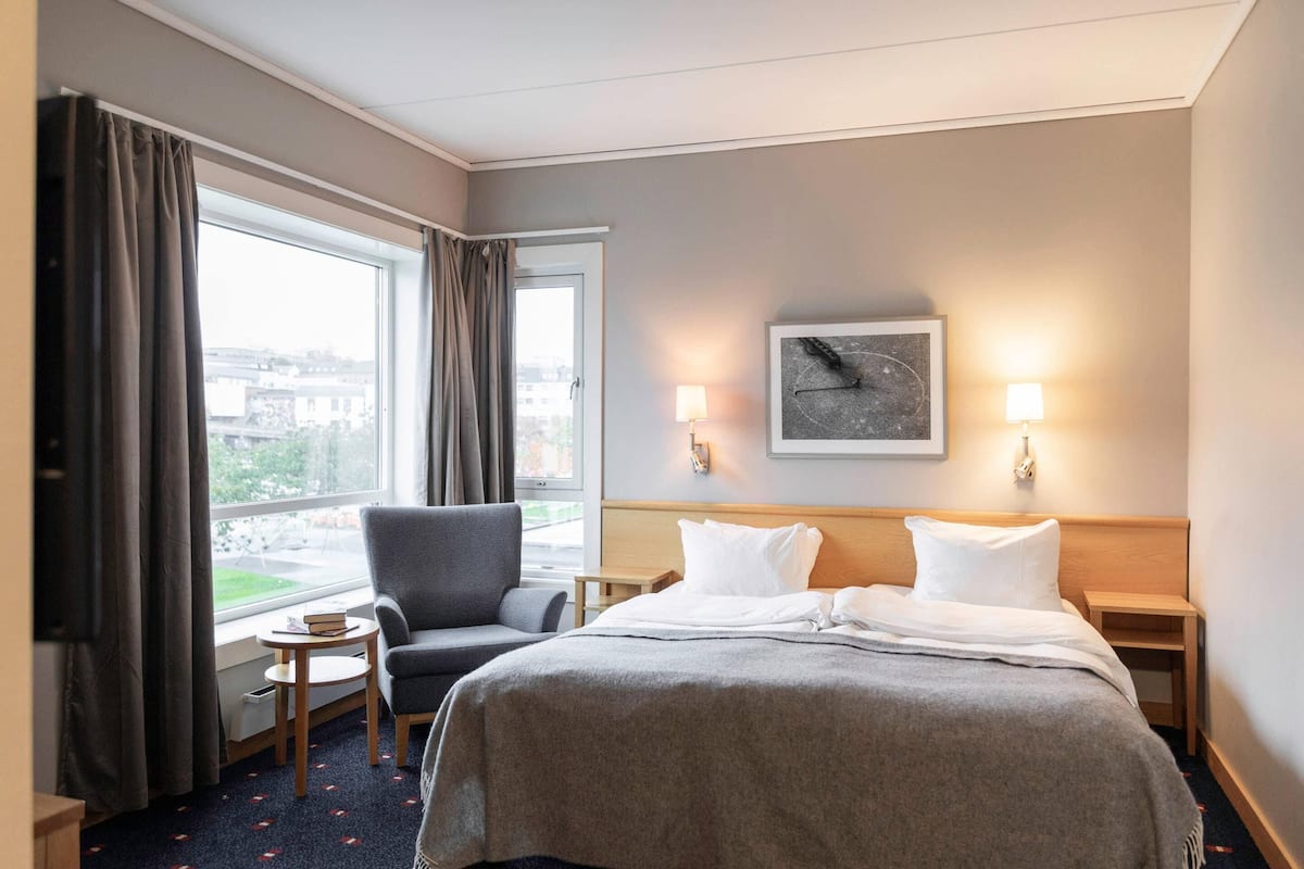 A hotel room with a bed, a chair, a small table, a lamp, and a window with curtains.