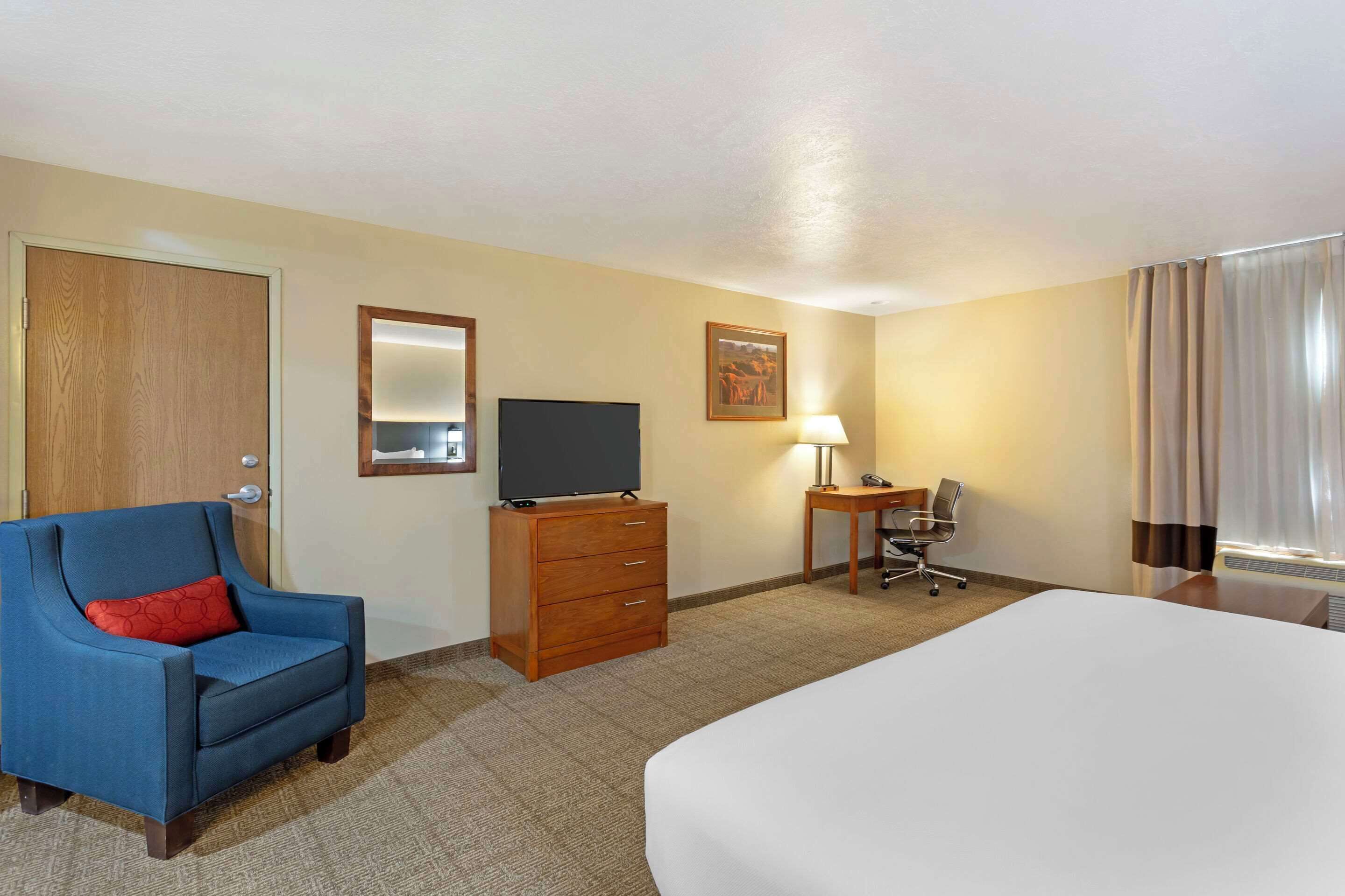 Une chambre d’hôtel avec un lit, un fauteuil bleu avec un coussin rouge, une commode en bois surmontée d’un téléviseur à écran plat, un bureau avec une lampe et une fenêtre avec des rideaux.