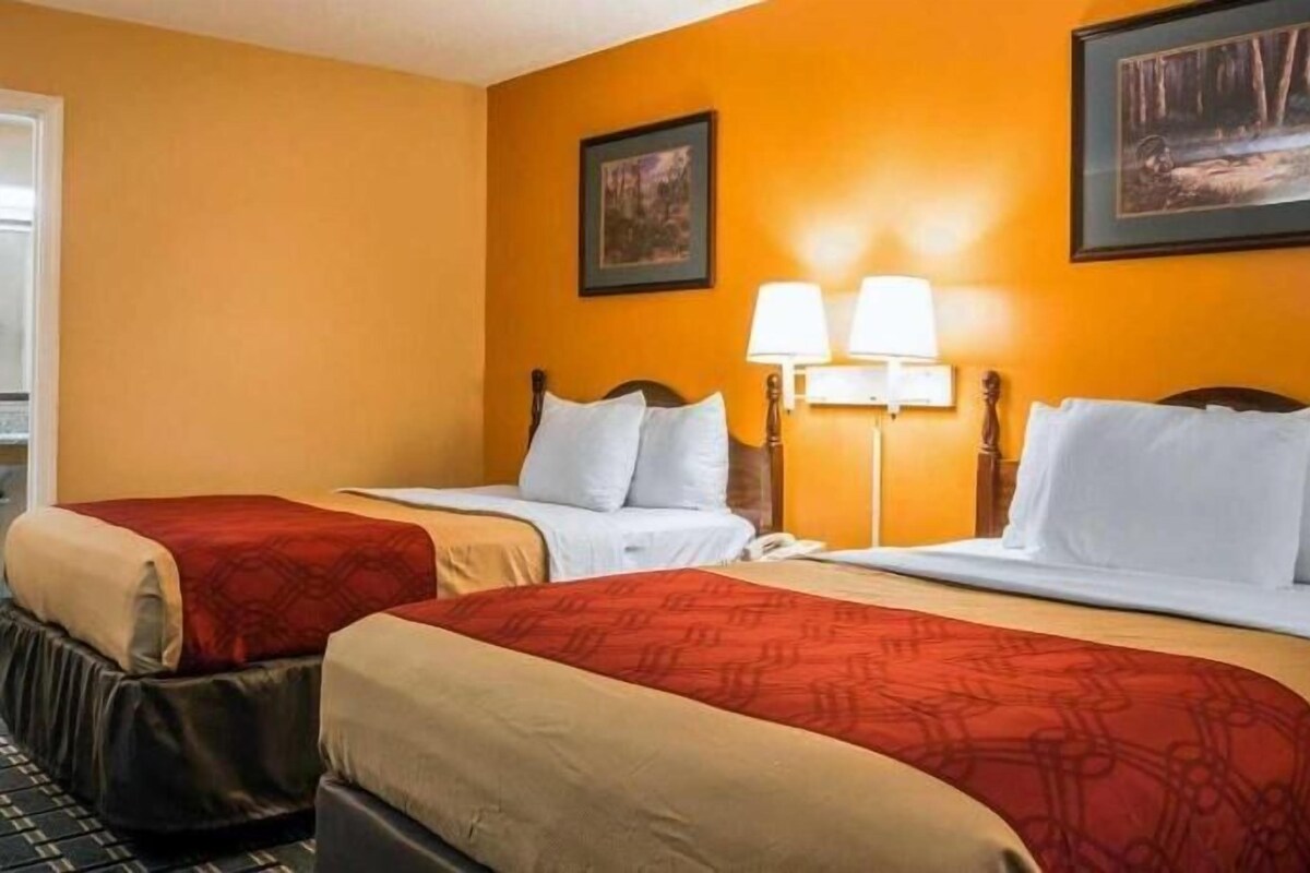 A hotel room with two beds, a red and beige bedspread, a wooden headboard, a nightstand, and two wall-mounted lamps.