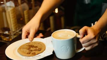 En person holder en kaffekop med latte-kunst, en tallerken med en cookie, og en Starbucks-kop.