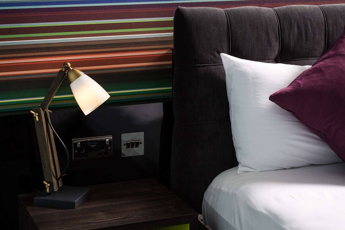 A bed with a white pillow and a purple pillow, a lamp on a wooden nightstand, and a striped wall.