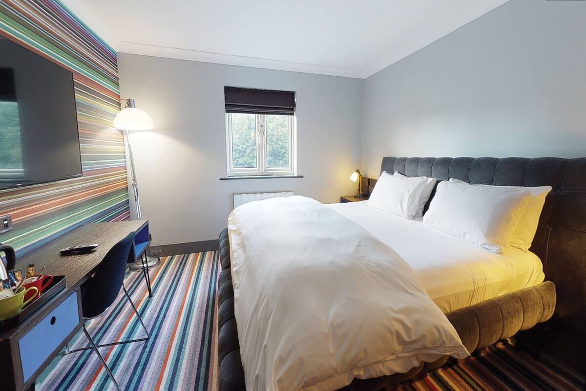 A hotel room with a large bed, a desk with a computer, a TV, and a window with blinds.