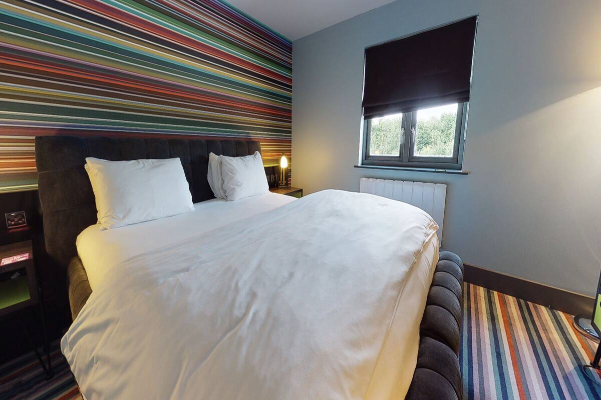 A modern hotel room with a large bed, a headboard with a built-in shelf, a window with a blackout blind, and a colorful striped wall.