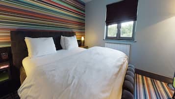 Quarto de hotel com uma cama grande, cabeceira, janela com persiana e parede listrada e colorida.