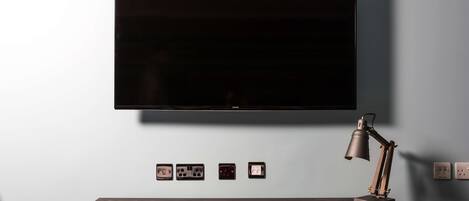 Una habitación de hotel moderna con un televisor de pantalla plana grande montado en la pared, un escritorio con una silla roja y una lámpara de escritorio.