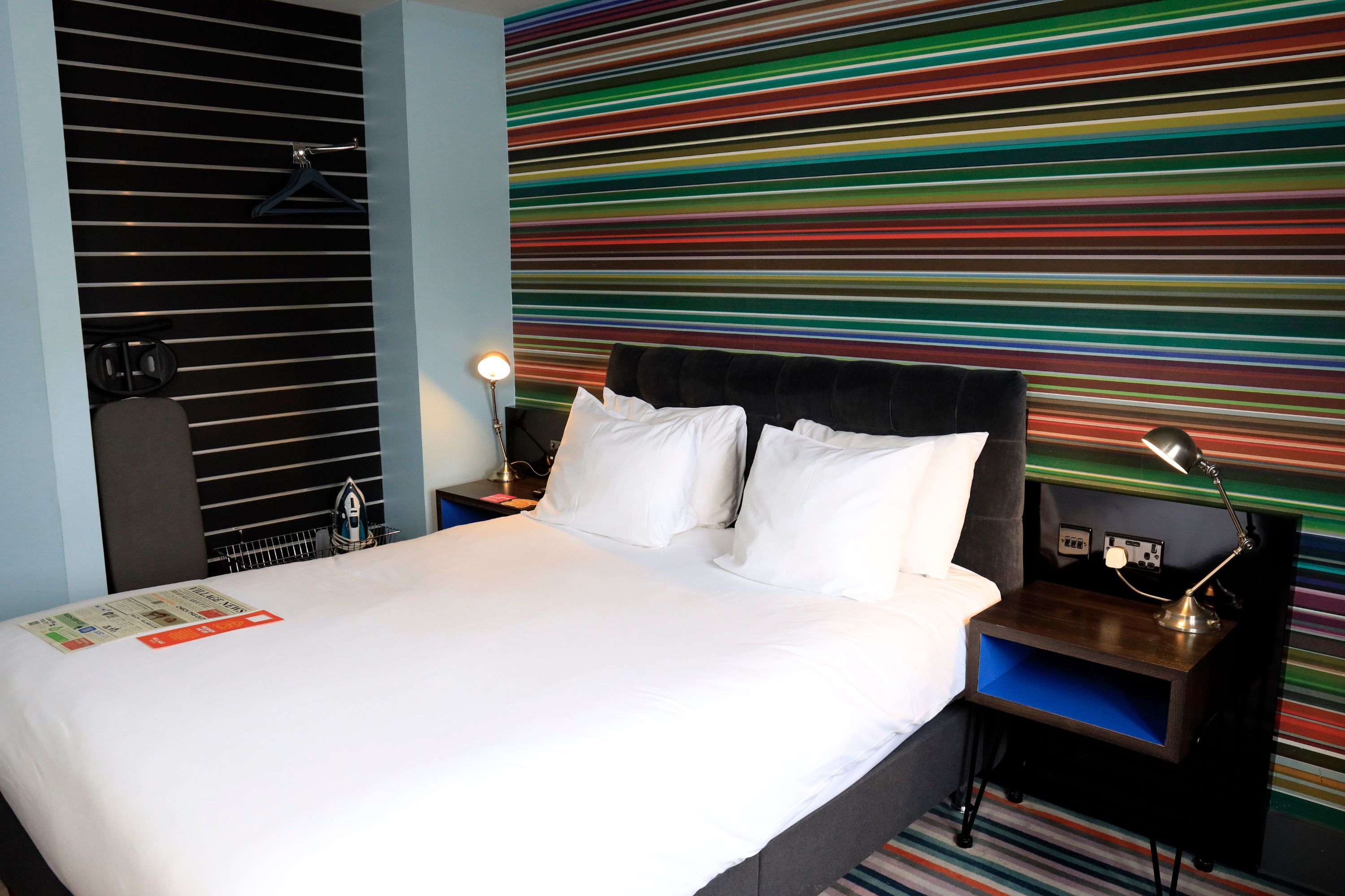 A hotel room with a bed, a nightstand, a lamp, a wall with horizontal stripes, and a window with blinds.