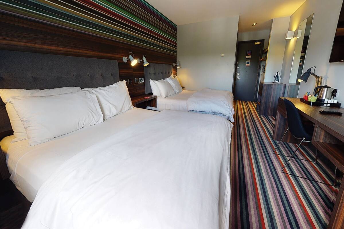 A hotel room with two beds, a desk, a chair, and a striped carpet.