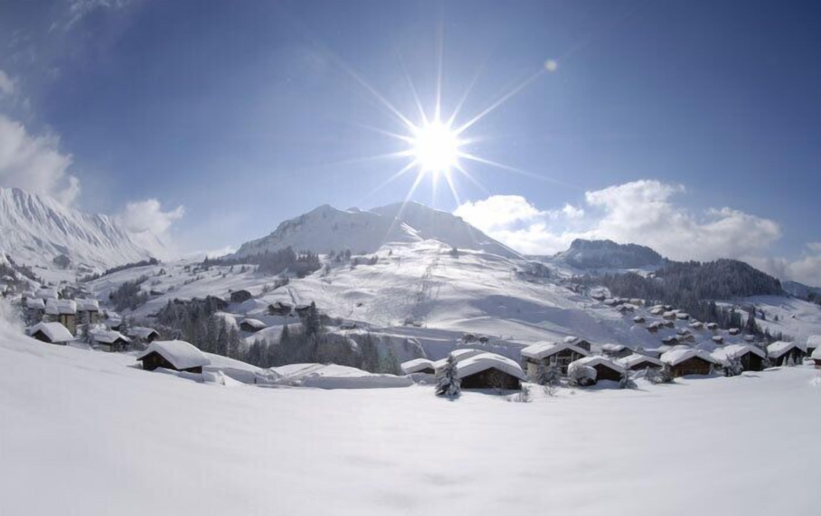 A snow-covered mountain village with traditional wooden houses, a clear sky, and a bright sun.