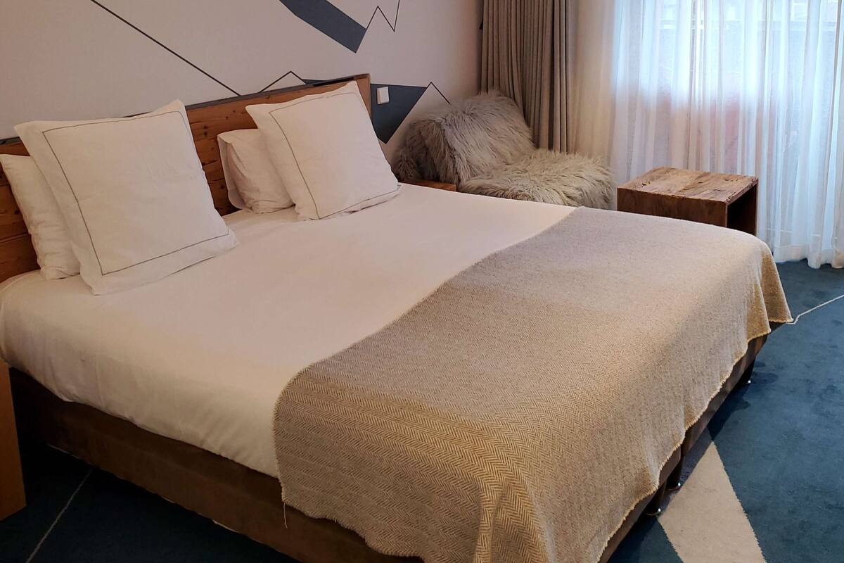 Habitació d'hotel amb un llit gran, una obra penjada a la paret, una tauleta de nit de fusta i vista a l'exterior a través de cortines primes.