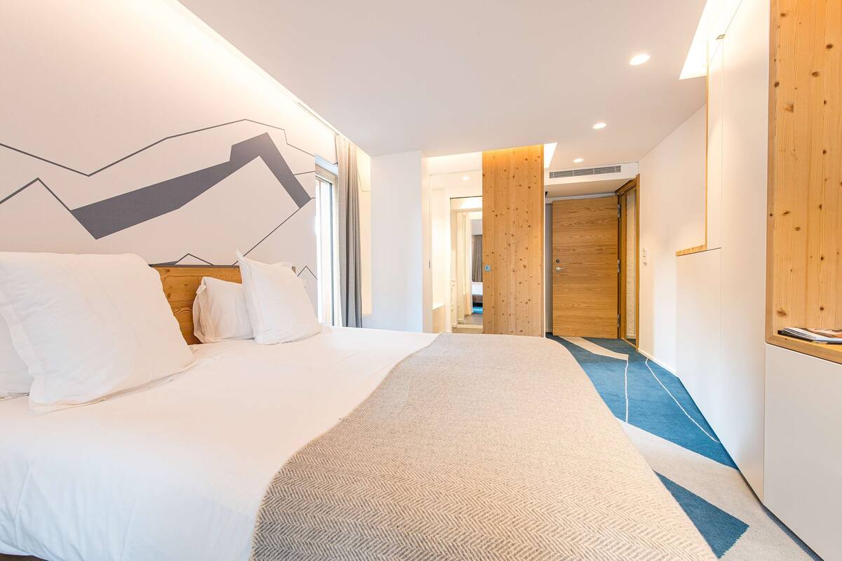 Una habitació d'hotel moderna amb un llit gran, un disseny de paret geomètric, detalls de fusta i un passadís que condueix a una altra habitació.