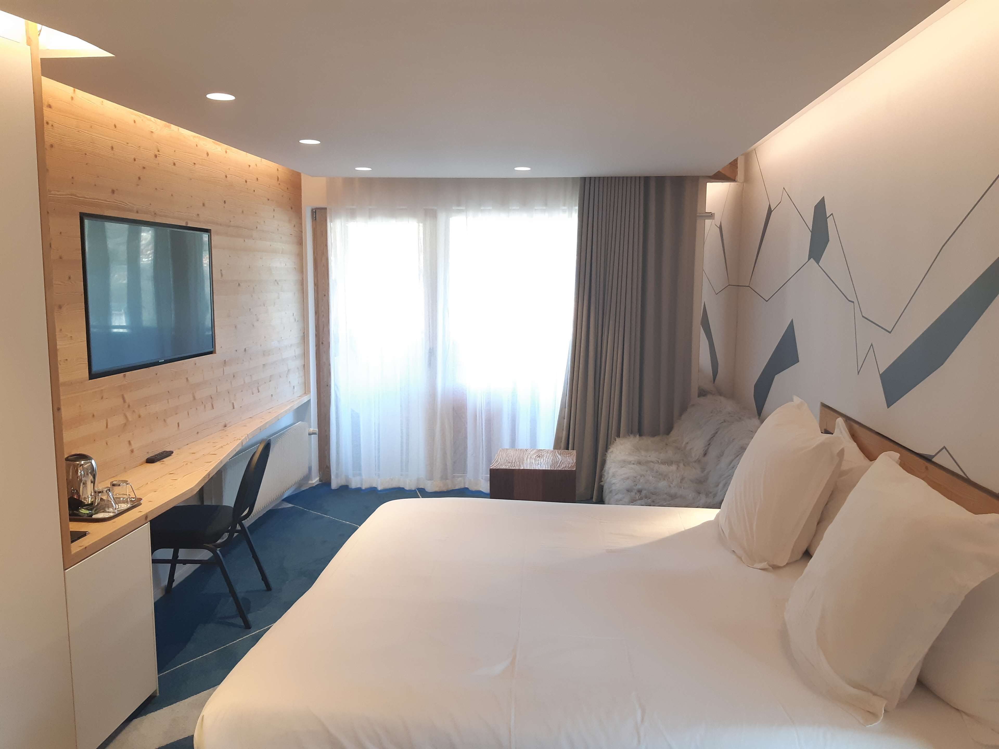 Ein Hotelzimmer mit einem großen Bett, einem Schreibtisch, einem Flachbildfernseher, einem Sessel und einem Fenster mit Vorhängen.
