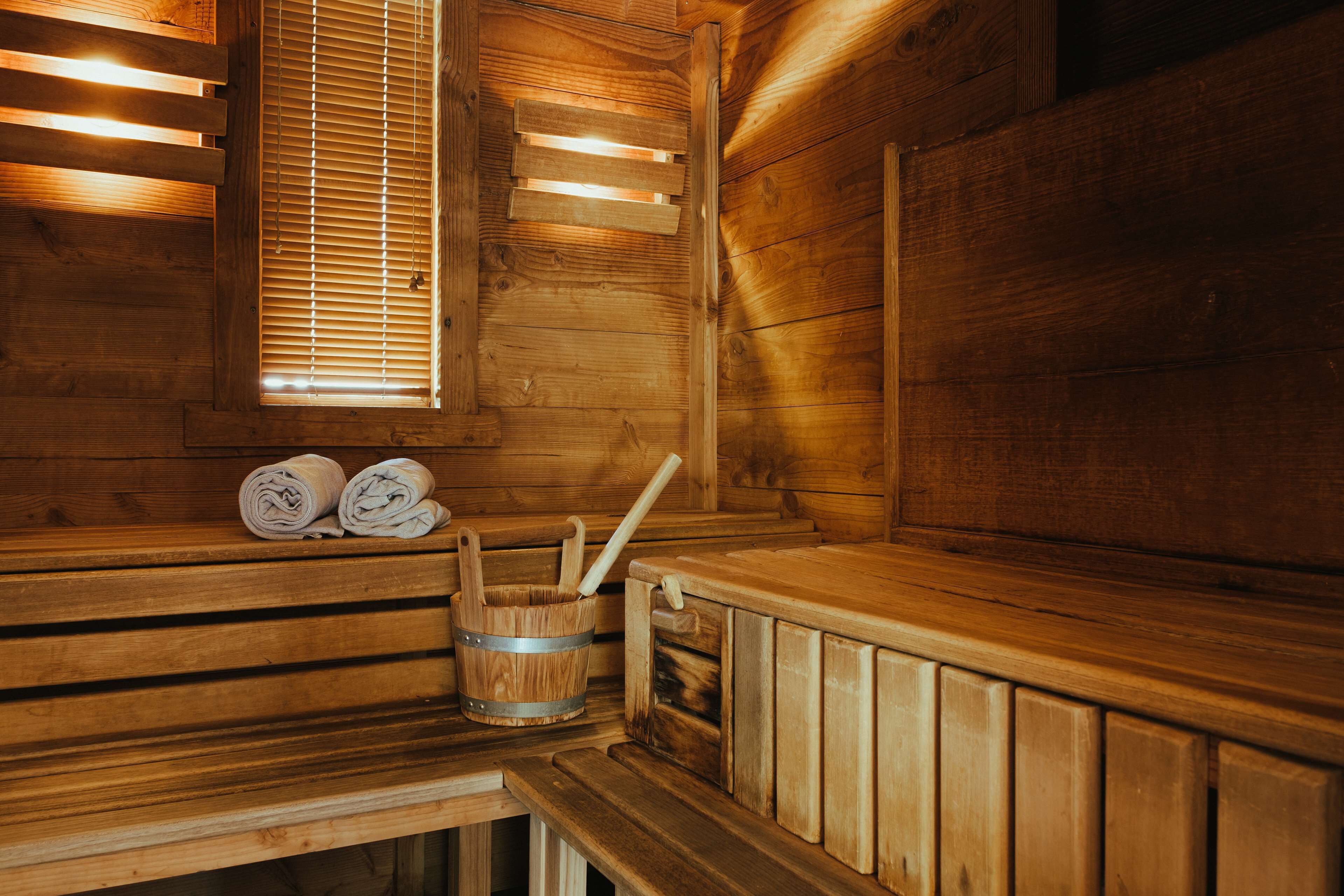 Una sauna de madera con bancos, una ventana con persianas y un cubo de madera con un cepillo.