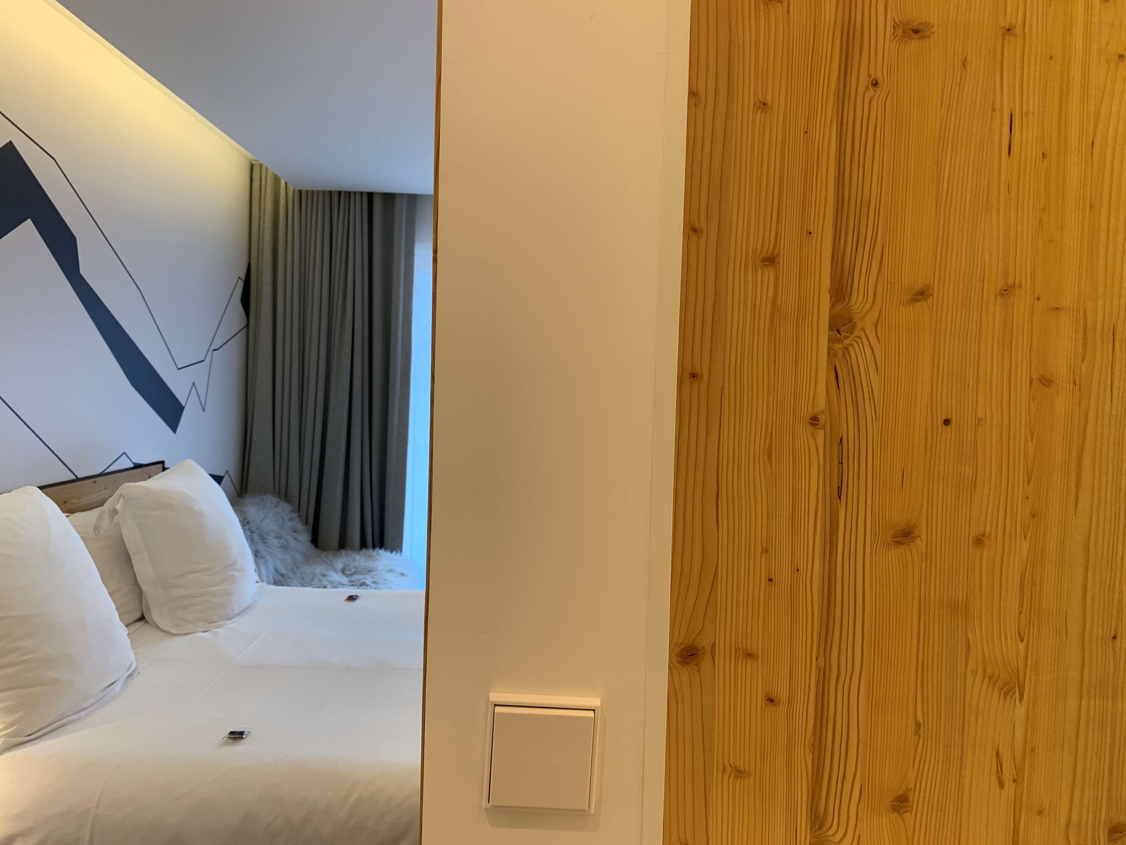 A bedroom with a bed, white bedding, a nightstand, a wall-mounted light, and a wooden door.