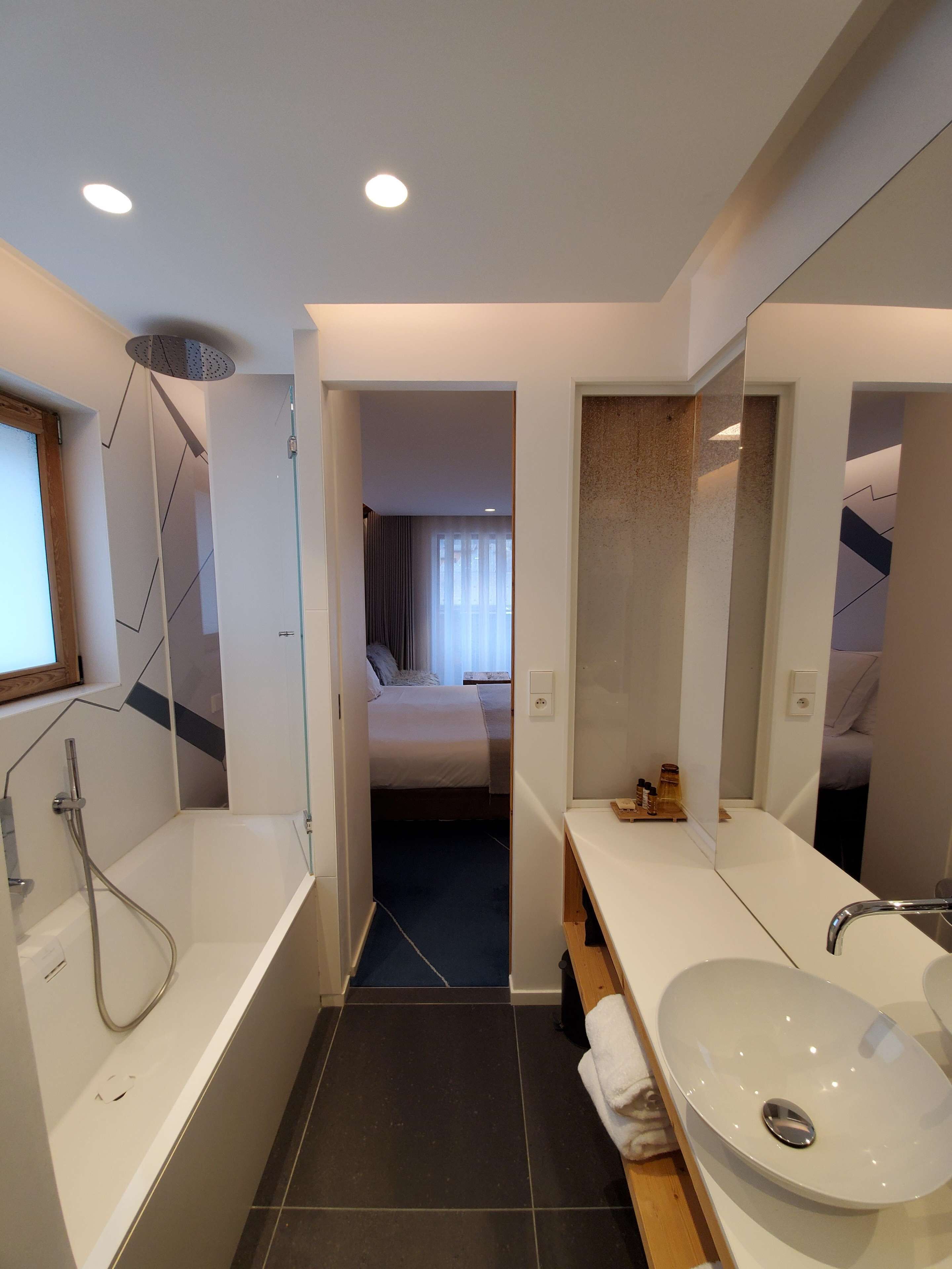 A modern bathroom with a bathtub, a sink, and a shower. There is a visible door leading to a bedroom.