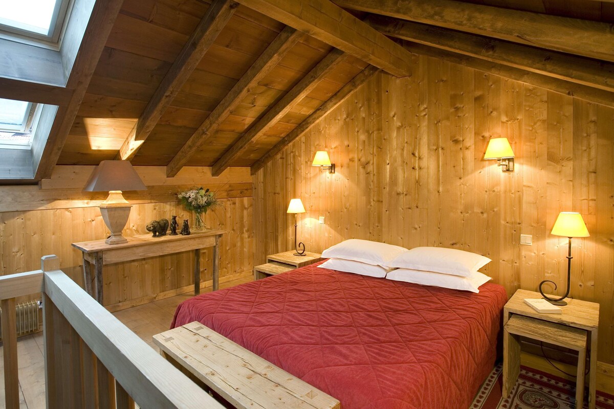 Una habitación acogedora de madera con una cama, dos mesitas de noche con lámparas, una pequeña mesa con un florero y un banco.