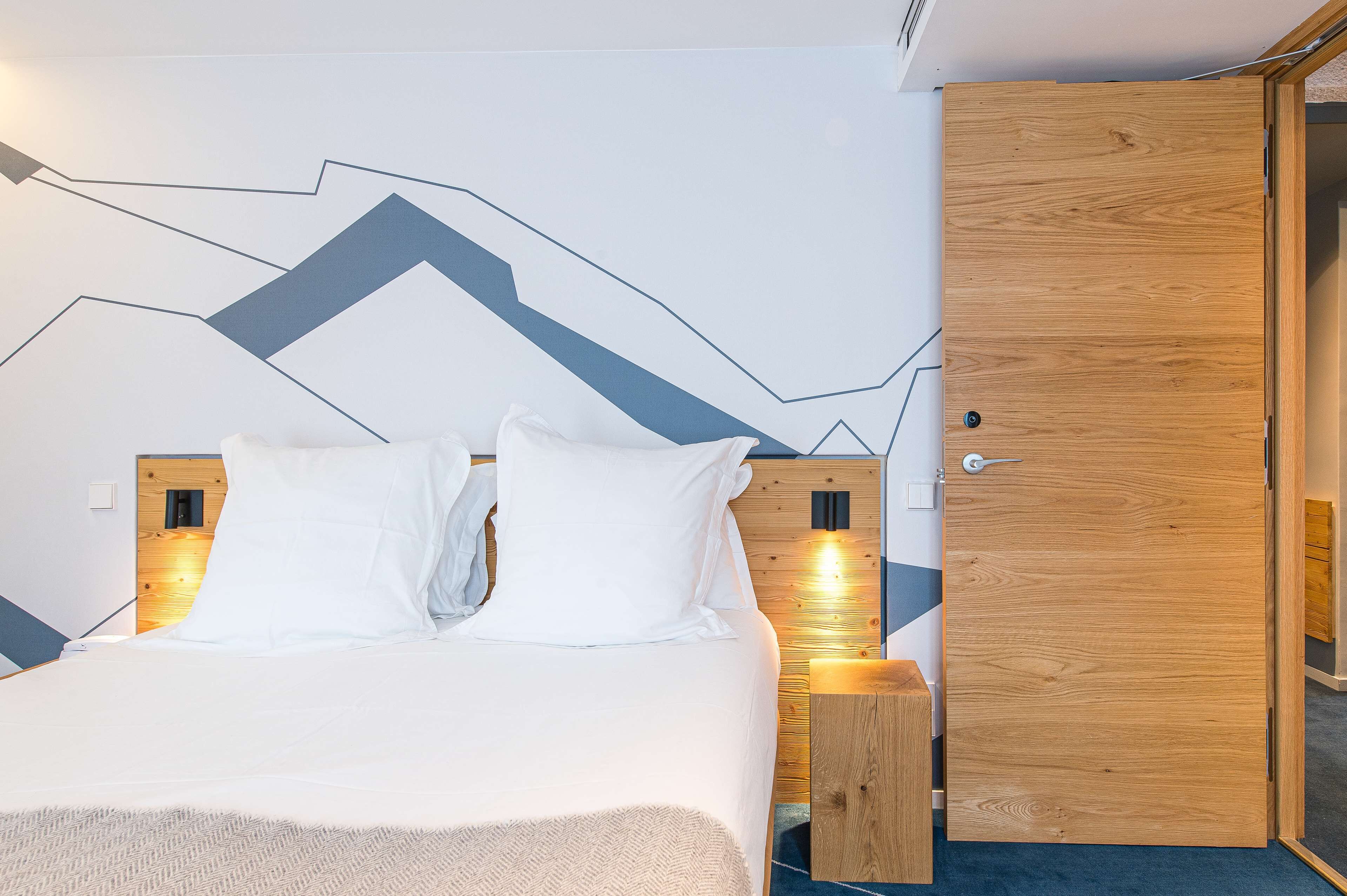 Una cama bien tendida con ropa blanca, cabecera de madera y una lámpara fijada a la pared.