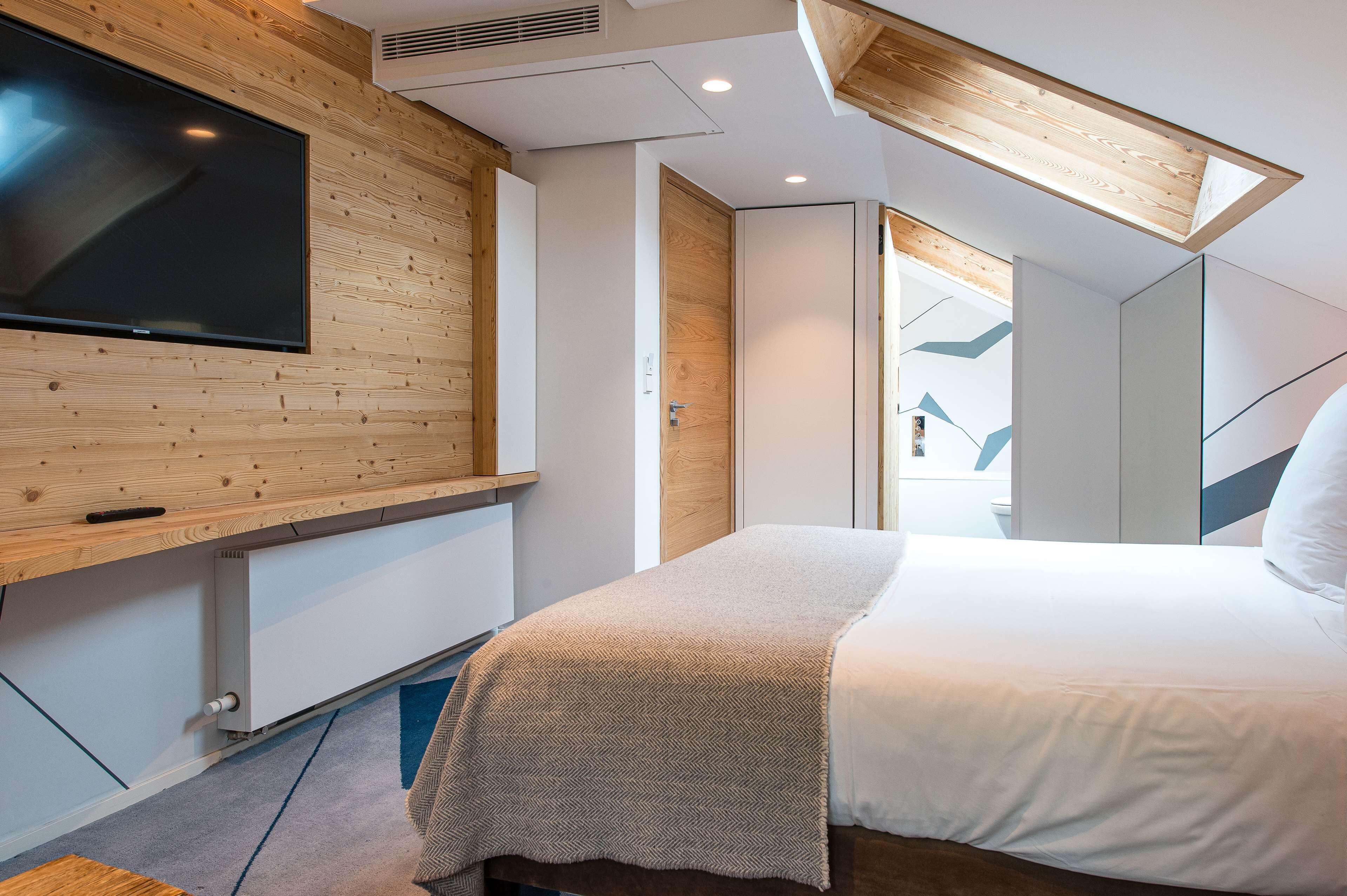 A modern bedroom with a large bed, a TV mounted on a wooden wall, and a skylight above the bed.