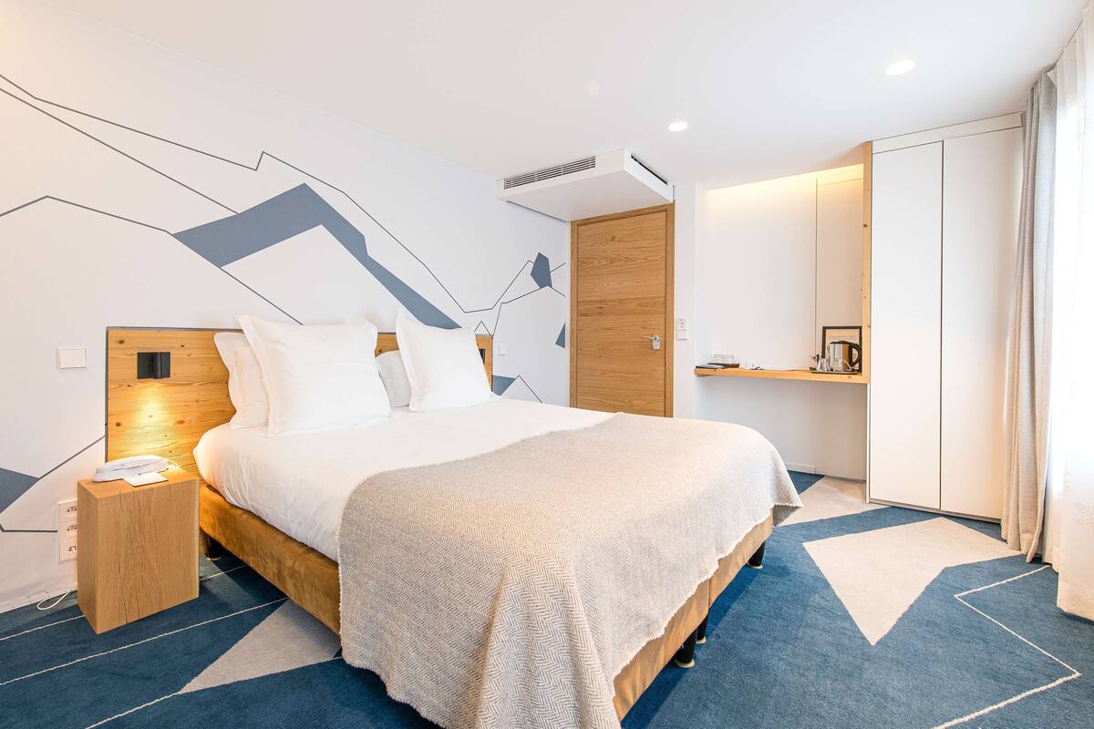Una habitació d'hotel moderna amb un llit gran, un panell de fusta al capçal, una taula de nit amb un telèfon i una catifes amb dibuixos geomètrics.
