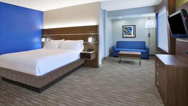 A hotel room with a large bed, a blue sofa, a small table, a TV, and a wooden cabinet. - Holiday Inn Express & Suites Manchester by IHG (Manchester)