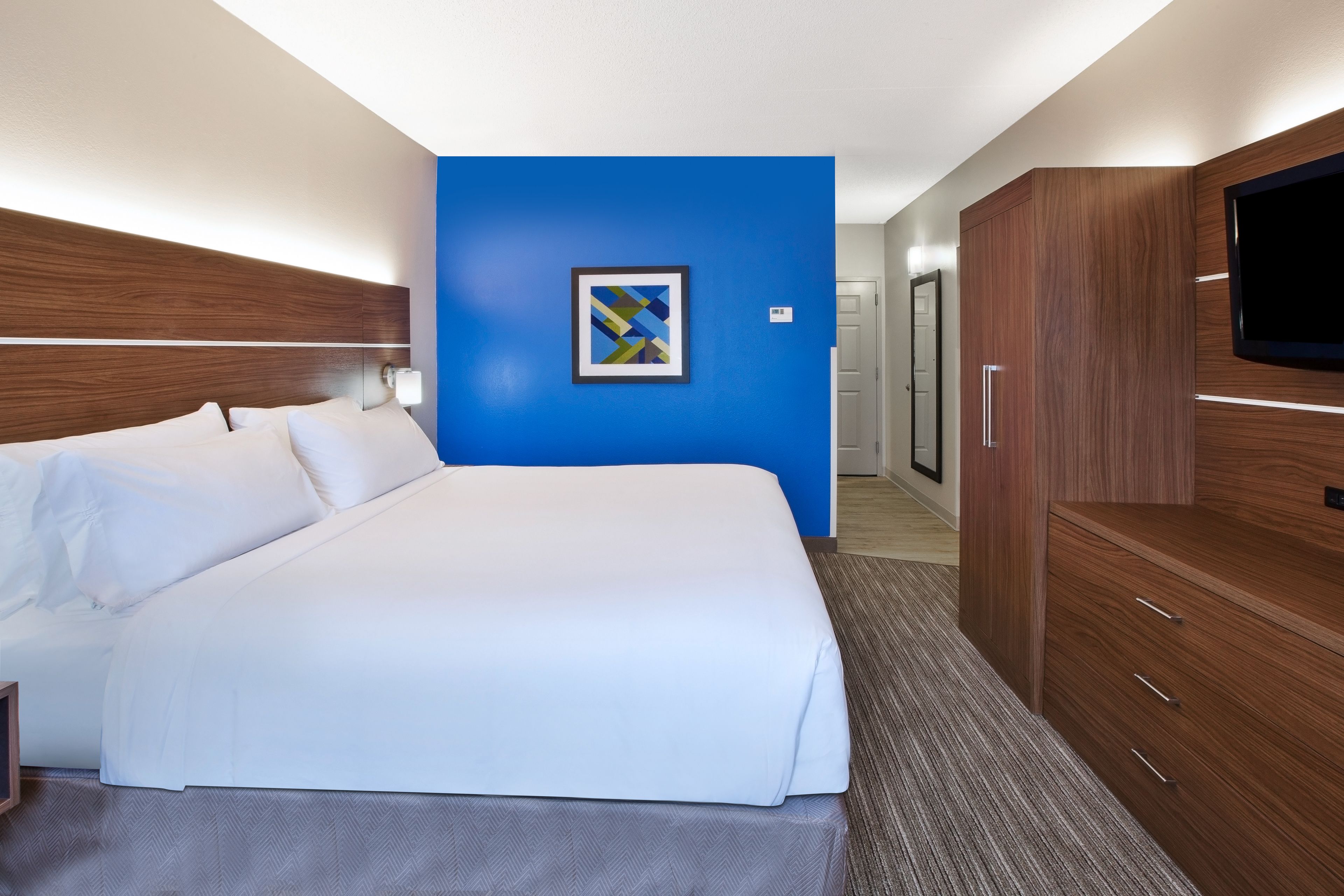 Quarto de hotel com uma cama grande, televisão, guarda-roupa e uma parede azul de destaque.