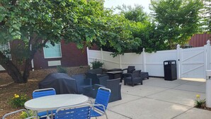An outdoor patio area with a round table and blue chairs, surrounded by dark wicker furniture, a covered grill, and a white fence. - Holiday Inn Express & Suites Manchester by IHG (Manchester)