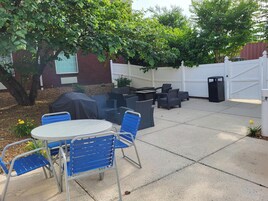 Un patio al aire libre con una mesa redonda y sillas azules, rodeado de muebles de mimbre oscuro, una parrilla cubierta y una valla blanca.