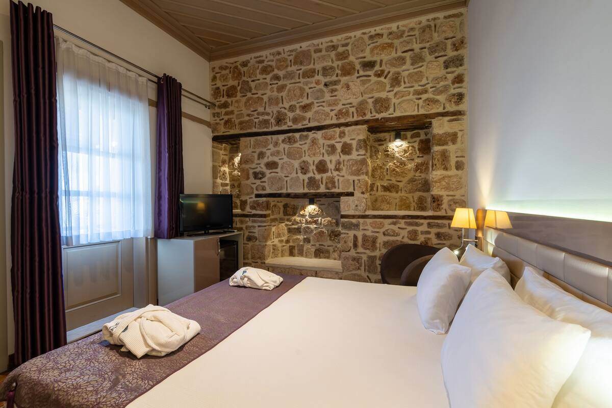 A hotel room with a stone wall, a bed with white linens, a TV on a stand, and a window with curtains.
