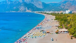 A coastal beach with rows of sunbeds and umbrellas, a clear blue sea, and mountains in the background. - Puding Hotel (Antalya)