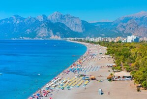 A coastal beach with rows of sunbeds and umbrellas, a clear blue sea, and mountains in the background.